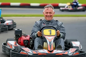 A middle aged man sitting on a go kart on an outdoor race track and he's smiling because he's racing for his 50th birthday party activity milestone