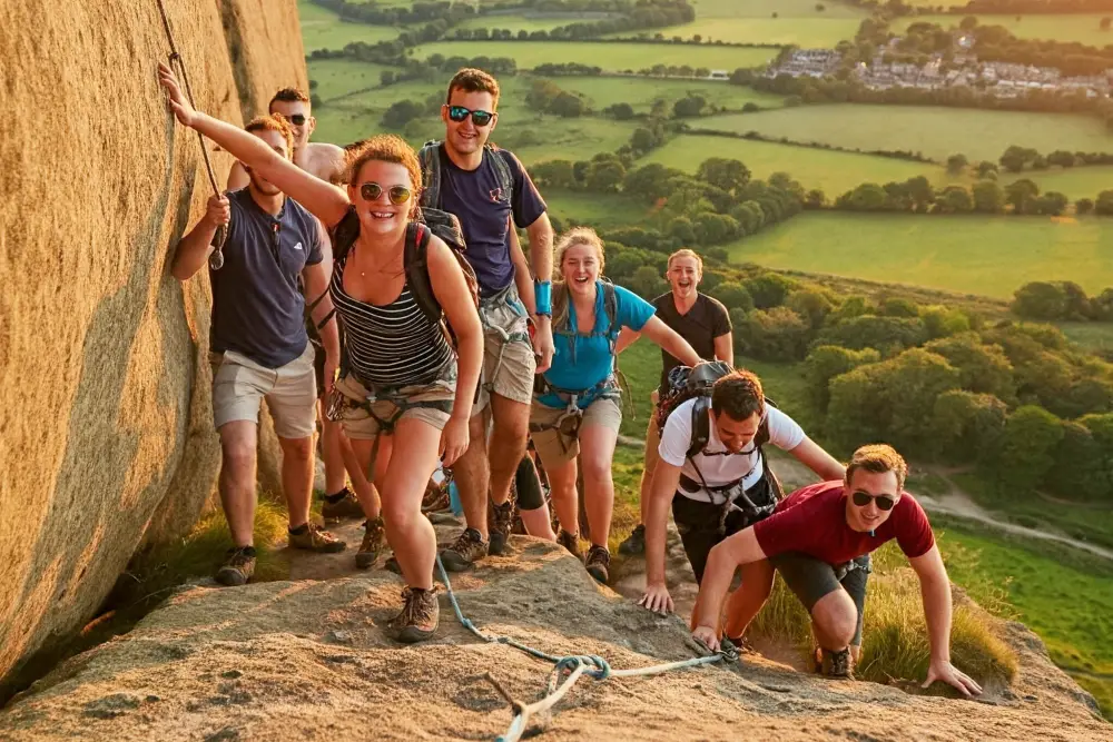 A group of adults rock climbing outside next to a cliff looking out over a green landscape for a group rock climbing activity