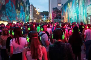 A large group of adults on a graffiti'd street and all wearing green neon headphones for a large silent disco walking tour at night time