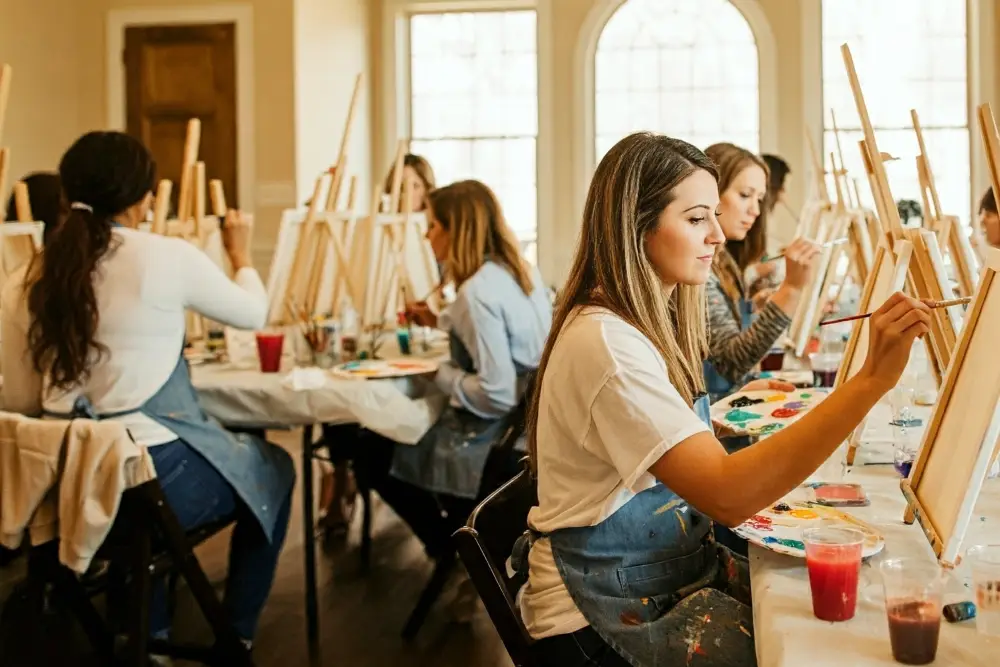 A group of adults in a paint and sip group activity class with canvases on easels, paints and glasses of wine on each table