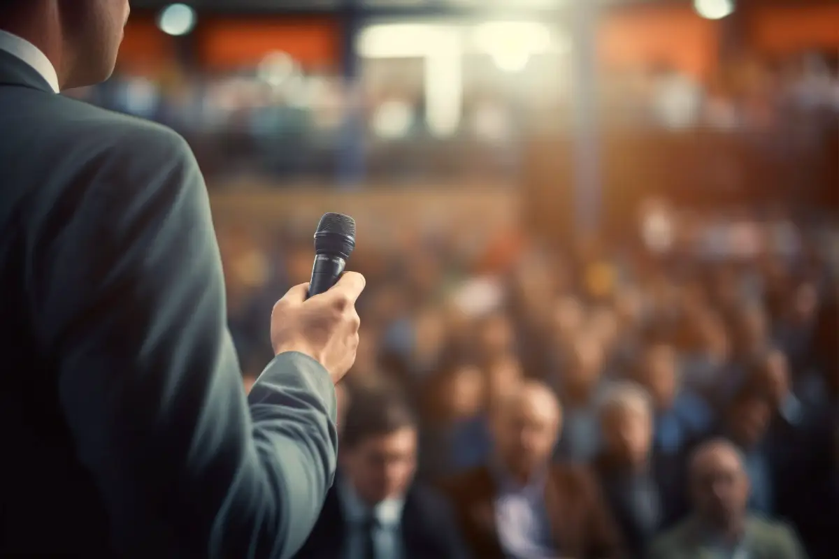 A man speaking to a large crowd through a microphone up on stage for a public speaking confidence workshop